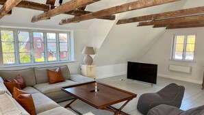 Un salón estilo loft con vigas de madera a la vista, un conjunto de sofás, una mesa de centro, un televisor y ventanas amplias con vistas a edificios.