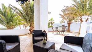 A patio area with wicker furniture, a table with a glass container, and a view of palm trees and a white wall. - Belvilla by OYO Holiday Home in Arico El Nuevo (Arico)