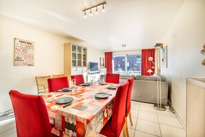 Un coin repas avec une table dressée pour six personnes, des chaises rouges, un meuble avec des portes en verre, un canapé et un radiateur.
