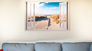 A framed beach scene with a wooden boardwalk and dunes, displayed above a grey sofa with a red pillow. - Apartment in Belgium Near the Sea (Koksijde)
