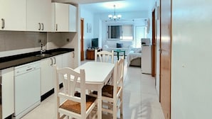 Una cocina compacta con armarios blancos y encimera negra, un comedor con sillas blancas, y una pequeña sala de estar con un sofá y un televisor.