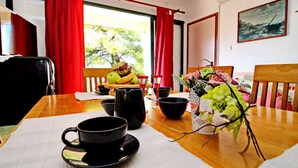 A dining table with a coffee cup, bowls, a pitcher, and a fruit bowl. There are red curtains, a window with a view of trees, and a painting on the wall. - Apartments Sunset - Two Bedroom Apartment With Terrace and Sea View (Blato)