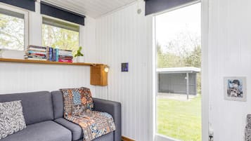 A cozy living room with a grey sofa, a patterned throw blanket, a bookshelf with books, and a large window with a view of the outdoors.