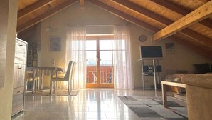 Amplia habitación en ático con techo de madera alto, puerta corrediza de vidrio que da a un balcón, televisor y sofá.