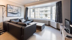Een moderne woonkamer met een bank, salontafel en een televisie. Er is een groot raam met gordijnen, een vloerlamp en een lijst met een afbeelding aan de muur.