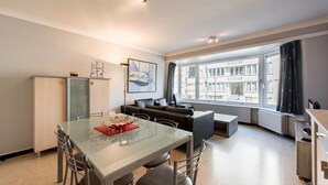 Een moderne woonkamer met een glazen eettafel, een bank en uitzicht op een gebouw door een groot raam.