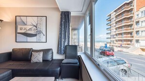 Een moderne hotelkamer met een zwarte leren bank, een klein tafeltje en een groot raam met uitzicht op een strand en een meer verdiepingen tellend gebouw.