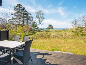 A deck with a table and chairs, overlooking a grassy area with trees and a view of the sea.