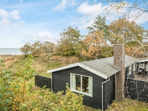 A small black cabin with a window, surrounded by natural vegetation and a view of the sea.