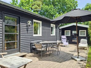 A wooden deck with a table and chairs, a patio umbrella, and a small building with windows. - 6 Person Holiday Home in Glesborg (Glesborg)