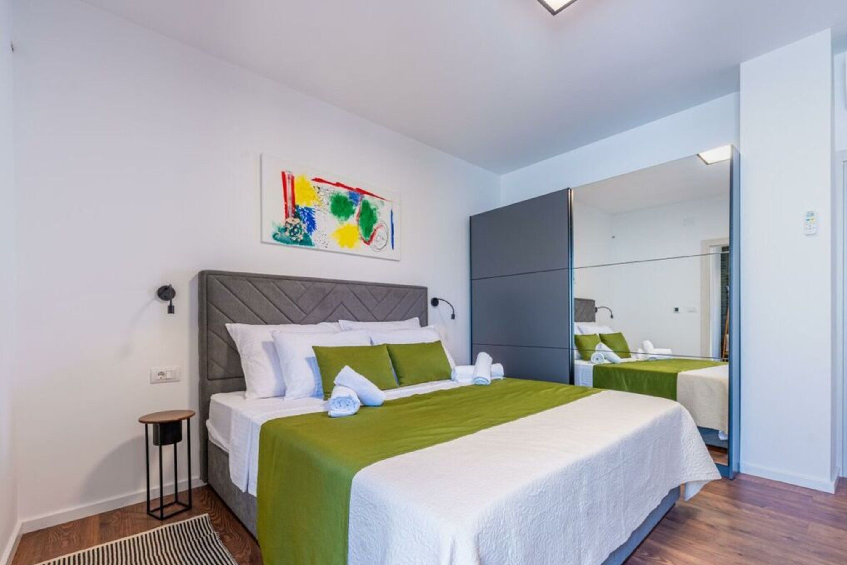 Habitación de hotel con una cama grande, una mesita de noche, un espejo y un cuadro abstracto de colores en la pared.