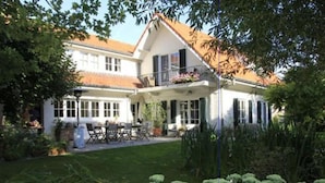 A two-story house with a balcony, a patio with outdoor furniture, and a well-maintained garden with white flowering plants. - Charming Home w/ Garden (Ostend)