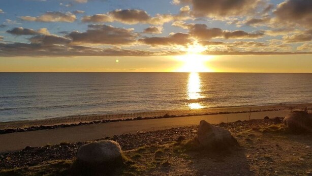 A sunset over a calm sea with a rocky shoreline and a paved path. - Siems Modern Retreat (Grube)