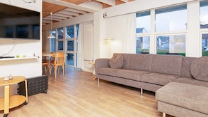 Ein modernes Wohnzimmer mit Holzboden, einem grauen Sofa, einem Essbereich mit Tisch und Stühlen sowie großen Fenstern mit Blick ins Freie.