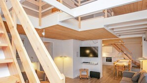 Ein geräumiges Loft mit hohen Decken, einer Holztreppe, einem an der Wand befestigten Flachbildfernseher, einem Essbereich mit Tisch und Stühlen sowie einer gemütlichen Sitzecke mit Sofa und Hocker.