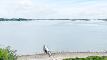 A coastal landscape with a pier extending into calm waters, surrounded by greenery and distant hills.