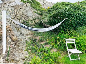 A hammock tied between rocks and a white chair in a garden with greenery and a stone wall.