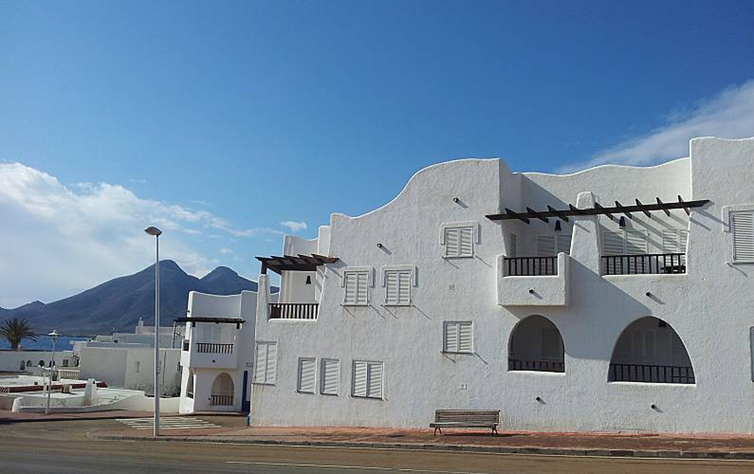 A white building with balconies and arched windows, a bench outside, and a mountain in the background.