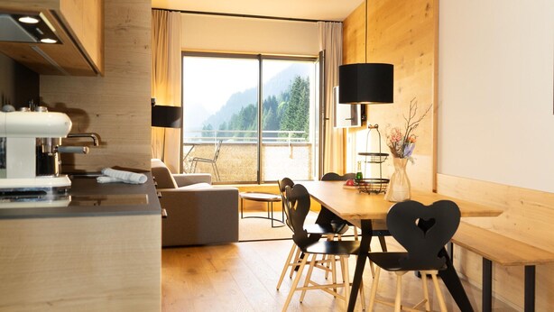 A modern kitchen with a coffee machine, a dining area with a wooden table and chairs, and a large window with a view of mountains. - Apartment in Schrocken Near Ski Arlberg (Schroecken)