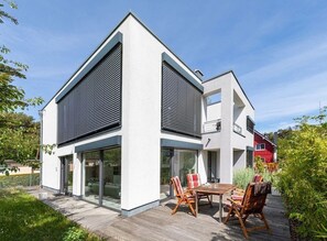 Una casa moderna de dos pisos amb grans finestres, una terrassa amb mobles d'exterior i un edifici vermell al fons.