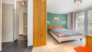 A modern bedroom with a bed, a wooden accent wall, a bathroom with a glass door, and a view of greenery outside. - Holiday Apartment Lore (Breege)