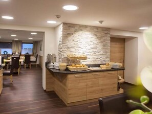 A modern kitchen with a stone accent wall, wooden cabinets, and a breakfast bar with bread and pastries. - Hotel Room in Langenfeld (Laengenfeld)