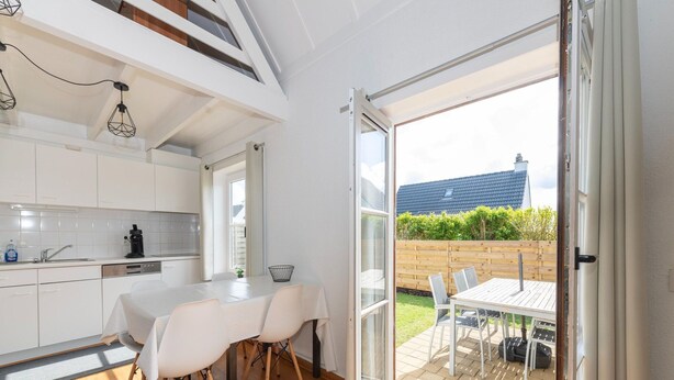Una cocina moderna con armarios blancos, una mesa de comedor con sillas y un ventanal corredizo que da a un patio exterior con terraza de madera y sillas.