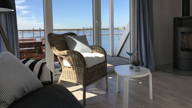 Une pièce avec un fauteuil en osier, une petite table blanche et une vue sur l’eau grâce à de grandes portes vitrées.