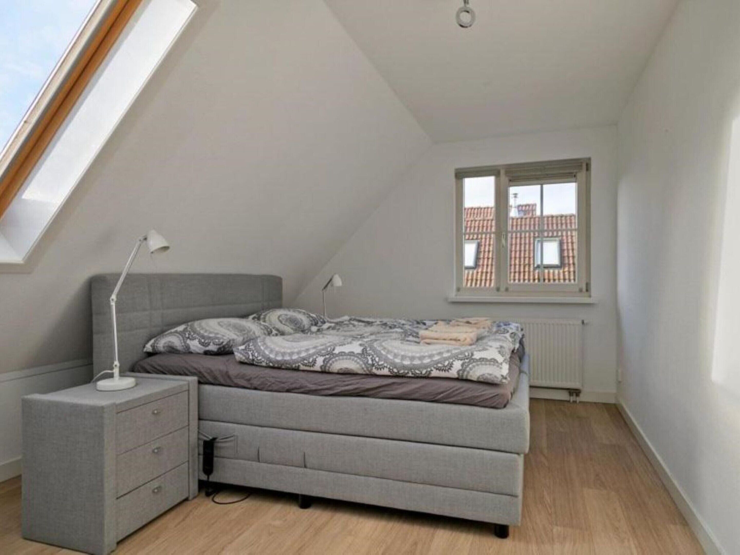Une chambre avec un plafond en pente, un lit avec une literie à motifs, une table de chevet avec une lampe et une fenêtre donnant sur des immeubles.