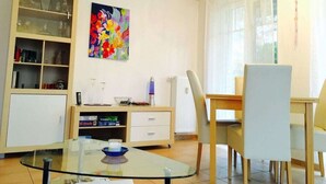 A modern living room with a glass coffee table, a white sofa, a dining table with chairs, a bookshelf, and a colorful abstract painting on the wall. - Holiday Apartment Sunny (Boltenhagen)
