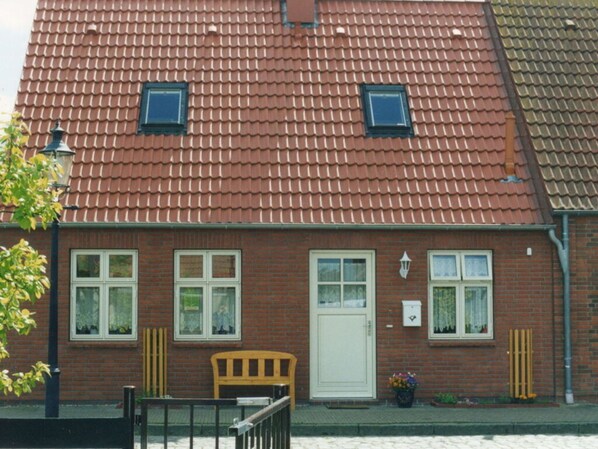 Een twee verdiepingen tellend, bakstenen huis met een rode tegelsdak, twee ramen met witte kaders, een witte deur en een kleine tuin met bloemen en een bankje.