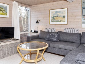 Ein Wohnzimmer mit grauem Sofa, einem Glastisch, einem Fernseher und gerahmten Kunstwerken an den Wänden.