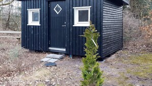 En liten, mörk trästuga med två fönster och en dörr, omgiven av träd och en träveranda.