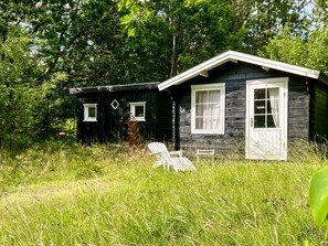 A small cabin with a single window and a door, surrounded by tall grass and trees. - 5 Person Holiday Home in Veslos (Vesløs)
