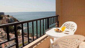 Un balcón con vistas al mar, una mesa con un plato de comida y un vaso de zumo de naranja, y sillas blancas.