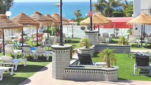 Zona de piscina en un complejo turístico, con sillones de descanso, sombrillas de paja y vista al océano.