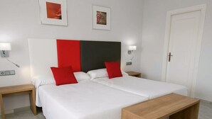 Habitación de hotel con dos camas, un banco de madera, ropa de cama blanca, almohadas rojas y dos cuadros en la pared.