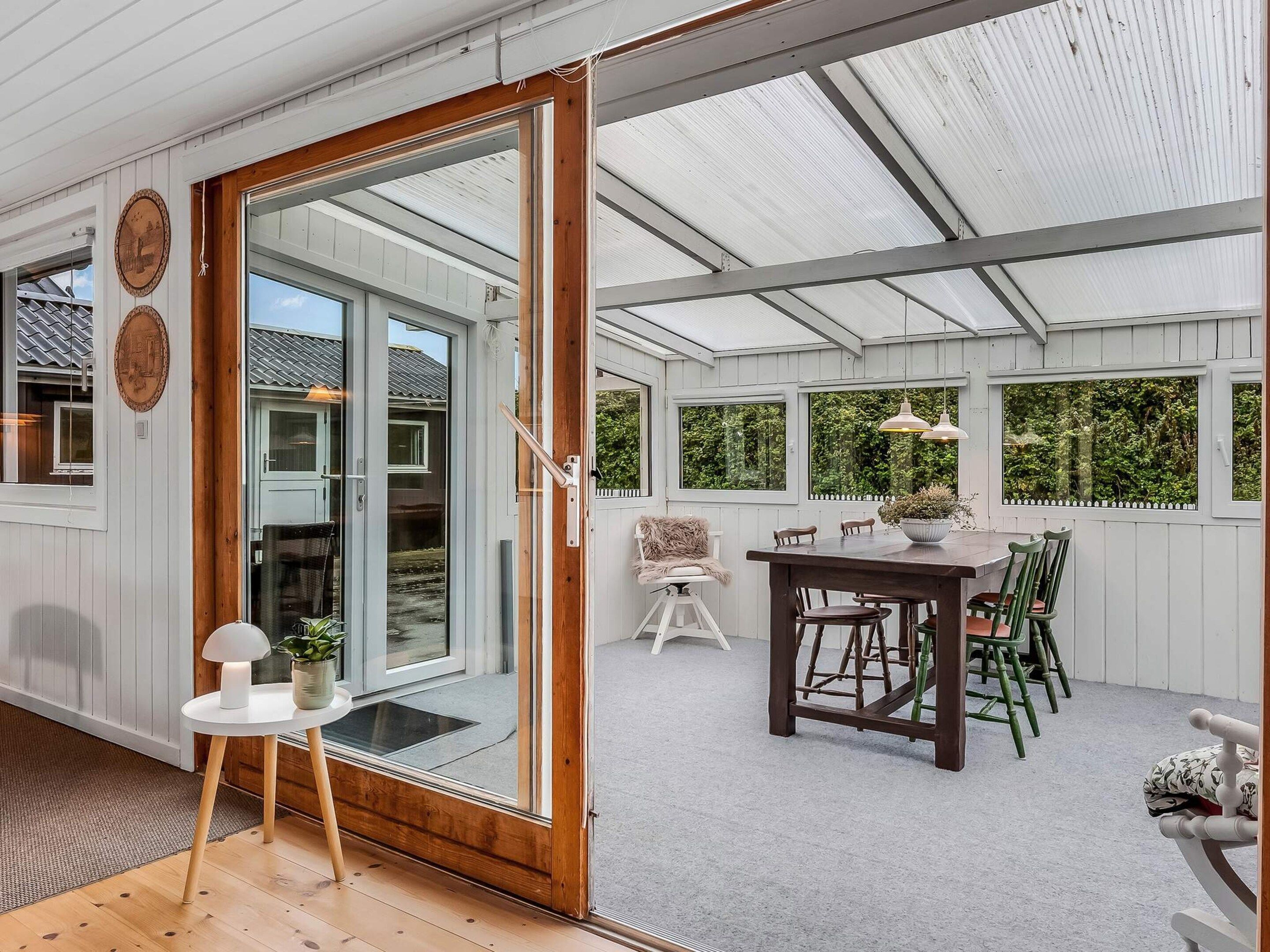 A covered patio area with a wooden table and chairs, a small side table with a lamp and plant, and a sliding glass door leading to an outdoor space.