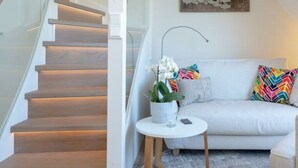 Ein modernes Wohnzimmer mit Treppe, einem weißen Sofa, farbenfrohen Kissen, einem kleinen runden Tisch mit Blumen und einem Wandbild.