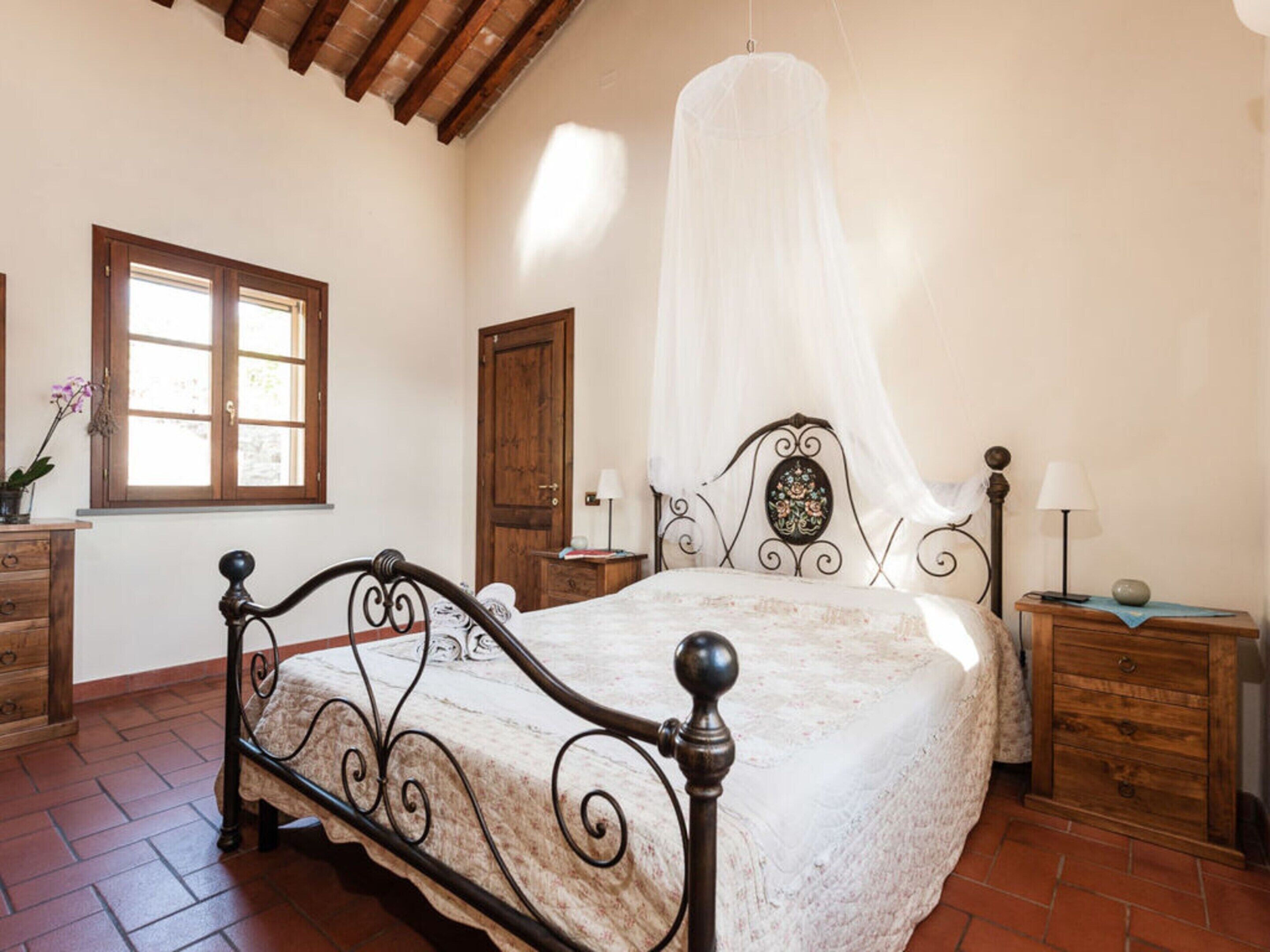 A bedroom with a four-poster bed, wooden headboard, and a canopy. There is a window with a view, a wooden dresser, and a nightstand with a lamp.