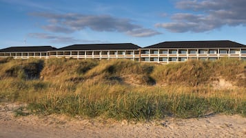 A modern building with multiple floors, situated on a sandy terrain with grass in the foreground.