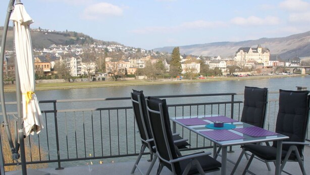 A balcony with a table and chairs overlooking a river, with buildings in the background. - Holiday Apartment "mosel & Umzu" (Bernkastel-Kues)