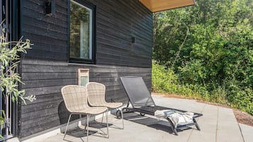 A modern house with a dark exterior, a large window, and a patio with two wicker chairs and a lounge chair.