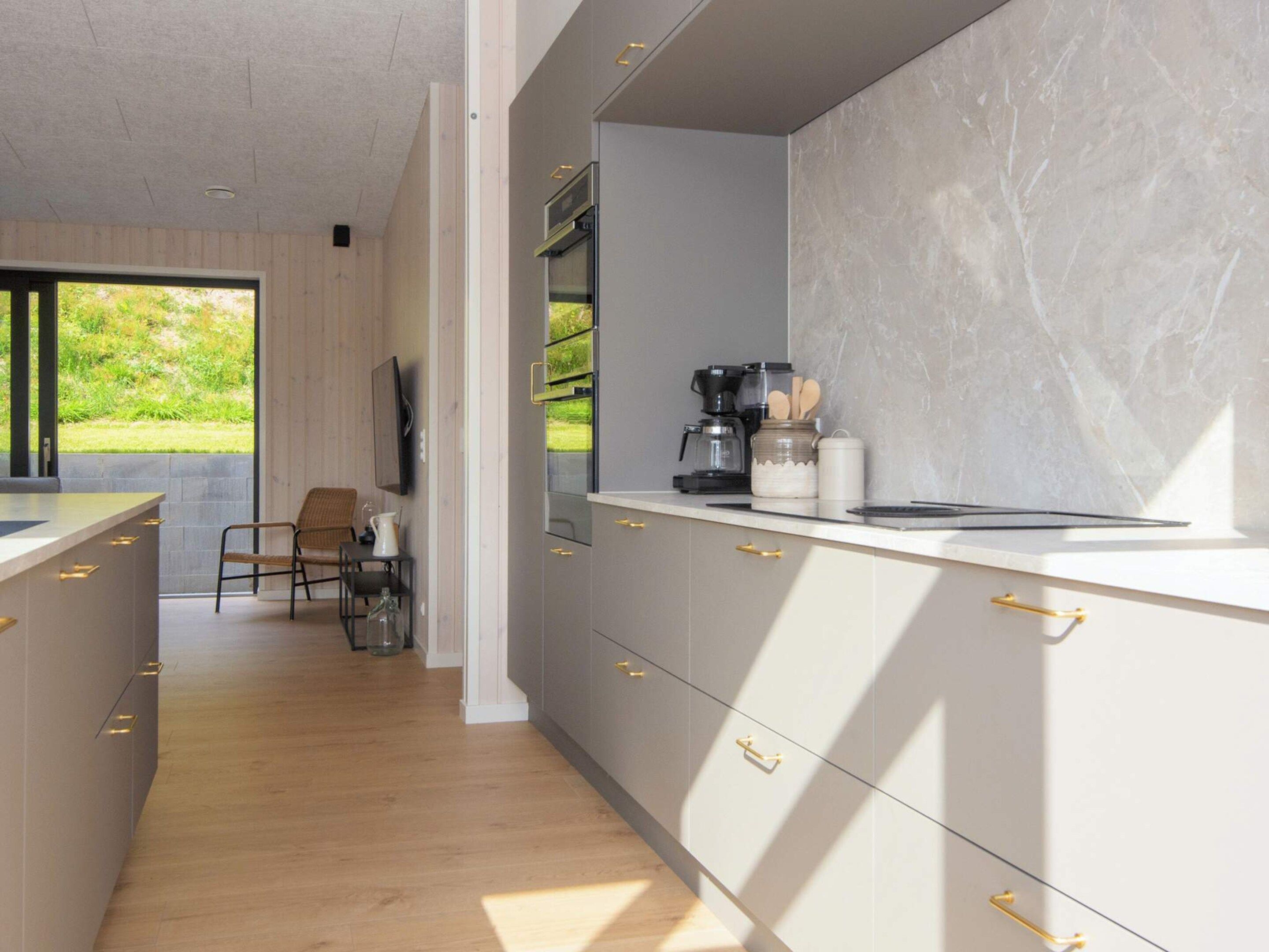 A modern kitchen with a marble backsplash, wooden flooring, and a view of greenery outside.