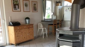 A cozy kitchen with a wood-burning stove, a wooden cabinet, and a dining area with a chair and table. - Holiday Home Steep Coast Fehmarn (Fehmarn)