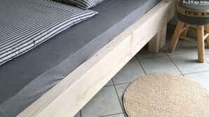 A bedroom with a wooden bed frame, grey bedding, striped pillows, a small stool, and a round rug on a tiled floor. - Holiday Home Steep Coast Fehmarn (Fehmarn)
