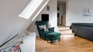 Ein modernes Loft-Apartment mit geneigter Decke, Holzboden, einem grünen Sessel, einer schwarzen Couch und einer Treppe zu einer erhöhten Ebene.