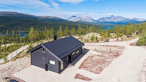 Una cabaña con techo oscuro, un pequeño porche y vistas a un lago y montañas.