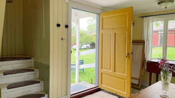 A room with a wooden staircase, a yellow door, a small table with a vase, and a view of a green lawn and parked cars outside.