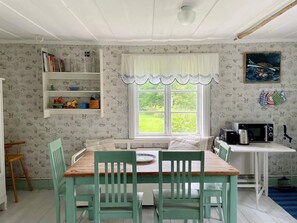 A dining area with a wooden table and green chairs, a window with a view of greenery, a shelf with books and toys, and a microwave on a white table. - 4 Person Holiday Home in Bjorko (Björkö)